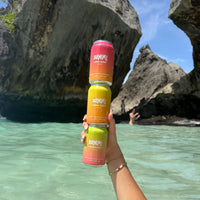 Hand holding three SUMMi Soda cans against a natural rock formation and water background